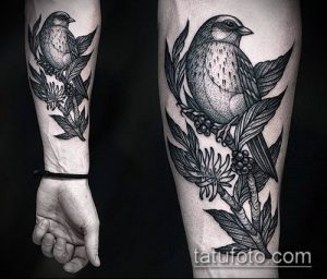 Фото татуировки птицы, сидящей на ветке с цветами. Черно-белый рисунок с детальной проработкой перьев и листьев. Эскиз тату на предплечье.