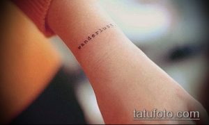 Фотография татуировки на запястье с надписью 'wanderlust' выполненной тонким шрифтом. Черно-белое изображение, минималистичный стиль.