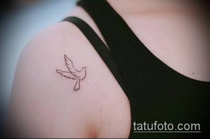 Черно-белое фото небольшой татуировки птицы в полете, расположенной на плече. Изображение выполнено тонкими линиями и выглядит очень изящно.