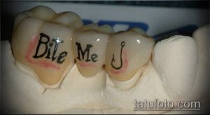 Фото татуировки на зубах с надписью 'Bite Me' и изображением рыболовного крючка. Рисунок выполнен в черном цвете.