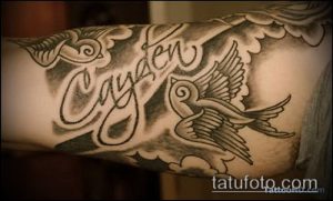 Фото тату двух ласточек, летящих вокруг надписи "Cayden" на руке. Черно-серые тона, детализированный рисунок татуировки, нежный стиль.