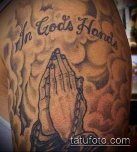 Фото тату молящихся рук на фоне облаков с надписью "In God's Hands". Черно-серые тона, религиозный рисунок татуировки, детальная проработка.