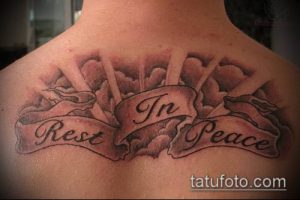 Фото тату на спине с надписью 'In Rest Peace' в облаках. Черно-серый рисунок татуировки с лентой и стилизованными облаками, символизирующими покой.