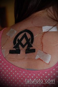 Фото свежей татуировки на спине, изображающей переплетенные буквы Альфа и Омега. Видны следы ухода и заживления.