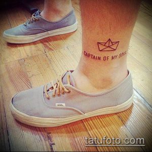 Фото тату оригами-лодки с надписью 'CAPTAIN OF MY SOUL' на лодыжке. Черно-белый рисунок татуировки в минималистичном стиле. Символичный и вдохновляющий дизайн.