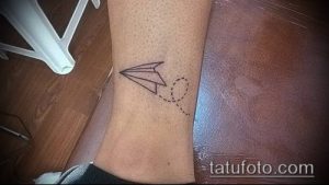 Небольшая черно-белая татуировка в стиле оригами на лодыжке. Изображен бумажный самолетик с пунктирной линией, символизирующей полет.