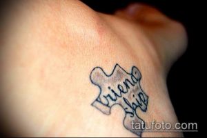 Фото тату на запястье с изображением элемента пазла, на котором написано слово "Friendship". Черный контур, простой дизайн.