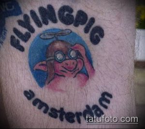 Фото тату летающего поросенка в голубом круге с надписью 'Flying Pig Amsterdam'. Рисунок выполнен в ярких цветах, с использованием черного контура.