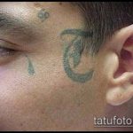 Фотография татуировки на лице мужчины: зеленый узор в стиле традиционной татуировки и капля слезы под глазом. Чёрно-белое изображение.