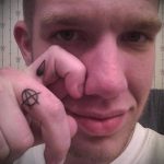 Фотография татуировки на пальце руки: маленький анархистский символ в круге. Черный рисунок на светлой коже.