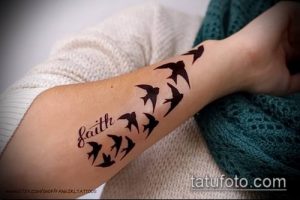 Фото тату нескольких черных ласточек в полете и надписи 'Faith' на предплечье. Элегантный и минималистичный рисунок татуировки.