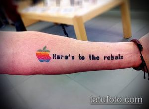 Фото татуировки с изображением логотипа Apple и надписью 'Here's to the rebels' на руке. Минималистичный рисунок татуировки для фанатов Apple.