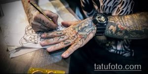 Фотография, демонстрирующая процесс создания эскиза татуировки. Мастер тату рисует карандашом на бумаге, а его рука с татуировками держит эскиз.