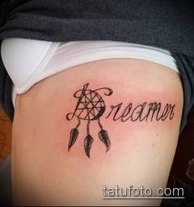 Фото татуировки с изображением ловца снов и надписи "Dreamer" на бедре девушки. Татуировка выполнена в коричневых тонах, с акцентом на детализацию перьев и плетения.