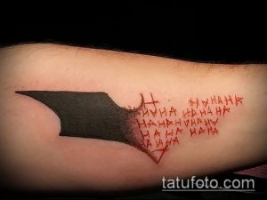 Фото татуировки с изображением символа Бэтмена, разрушающегося и превращающегося в надпись "HAHAHA" в стиле комиксов. Татуировка выполнена в черно-красной гамме.