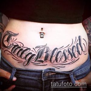 Фото татуировки на животе с надписью "Tracy Blessed", выполненной в стиле Blackwork. Рисунок выполнен черными чернилами с использованием плотных штрихов.