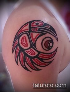 Фото татуировки в стиле Haida Raven на плече, с изображением головы птицы в круге, выполненной в черном и красном цветах.