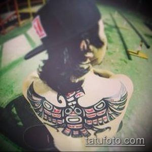 Фото татуировки в стиле Haida Raven на лопатке девушки, с изображением птицы, выполненной в черном и красном цветах.