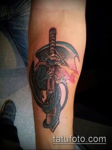 Фото тату на предплечье: дракон обвивается вокруг меча, из пасти дракона вырывается пламя, выполненное в ярких цветах с использованием теней.