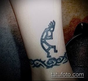 Черно-белая татуировка Кокопелли на запястье, окруженная трайбл-узором. Сочетание традиционного символа и современного стиля.
