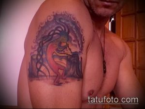 Фото тату Кокопелли на плече. Цветная татуировка в стиле этнической графики, изображающая танцующего духа на фоне стилизованных облаков и гор.