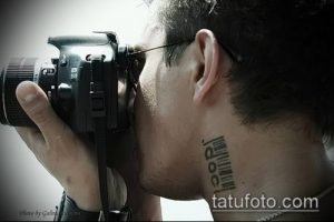 Крупный план шеи с татуировкой штрих-кода и фотокамерой в руках. Черно-белое фото демонстрирует современный рисунок татуировки.