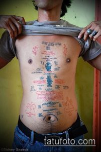 Фото татуировки, покрывающей большую часть тела. Изображены различные символы, надписи на разных языках и рисунки, выполненные в черном и красном цветах.