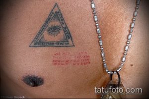 Фото татуировки на груди с изображением треугольника с глазом внутри и надписью. Рисунок выполнен в черно-белой гамме.