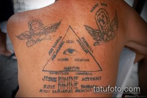 Фото татуировки на спине с изображением треугольника, ангелов и надписей на латыни. Рисунок выполнен в черно-белой гамме.