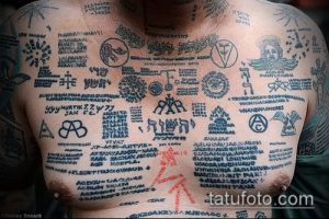 Фото татуировки, покрывающей большую часть груди и плеч. Изображены различные символы, надписи на разных языках и рисунки, выполненные в черном и красном цветах.