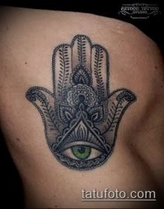 Фото тату Хамса (Рука Фатимы) на боку. Рисунок выполнен в технике blackwork с использованием сложных узоров и зеленого глаза.