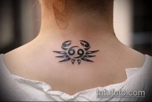 Фото тату: маленький черный рисунок знака зодиака Рак на спине девушки. Эскиз выполнен в минималистичном стиле с четкими линиями.
