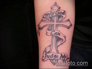 Фото тату надписи 'Only God Can Judge Me' с изображением креста и ленты на руке. Черно-серый рисунок татуировки с религиозной символикой.