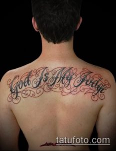 Фото тату надписи 'God Is My Judge' на спине. Изящный шрифт с завитушками и детальной проработкой коричневым цветом.