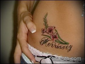Фото тату: небольшой гладиолус с надписью "Morrissey" на боку. Простой и элегантный рисунок татуировки.