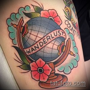 Цветная татуировка глобуса в стиле олдскул на предплечье, с надписью "Wanderlust" на ленте, опоясывающей глобус. Рисунок украшен цветами и облаками.