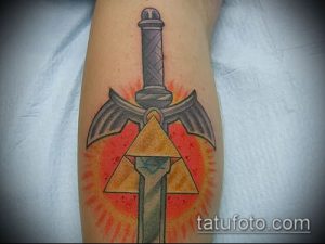 Фото тату на предплечье с изображением меча и трифорса из игры Zelda. Яркий рисунок татуировки в стиле фэнтези с использованием оранжевого и серого цветов.