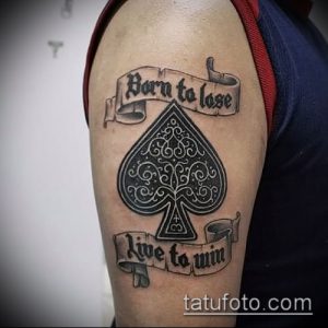Фото татуировки на плече: стилизованный туз пик с надписью 'Born to Lose, Live to Win' в виде лент.
