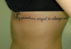 Фото татуировки надписи "My guardian angel is always with me" на боку, выполненной каллиграфическим шрифтом.