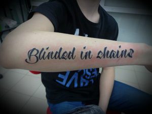 Фотография текстовой татуировки с надписью "Blinded in chains" на предплечье. Шрифт выполнен в каллиграфическом стиле.