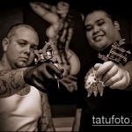 Фотография двух мастеров тату в студии, один держит тату-машинку, другой демонстрирует готовый эскиз. Черно-белое изображение, профессиональная татуировка.