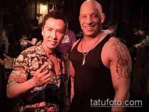 Фотография Вин Дизеля с татуировками на руках, улыбающегося и позирующего для камеры. На заднем плане видны люди и интерьер помещения.