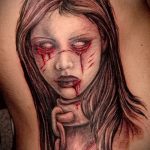 Черно-красная татуировка на руке, изображающая лицо плачущей девушки с кровью, стекающей по щекам.