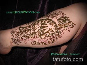 Фото тату: яркая татуировка Чеширского кота из "Алисы в Стране чудес" с чашкой чая. Рисунок в технике хендпоук на руке.