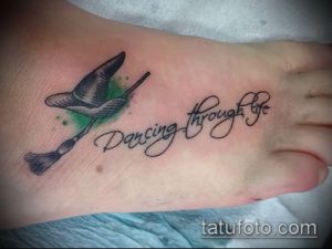 Фото тату на стопе: метла с надписью 'Dancing Through Life' выполнена в черно-серых тонах с зеленым акцентом.
