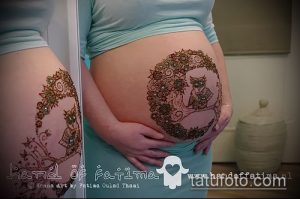 Фото мехенди на животе беременной женщины, изображающее сову, сидящую в центре цветочного венка. Рисунок выполнен в коричневых тонах с использованием сложных узоров.