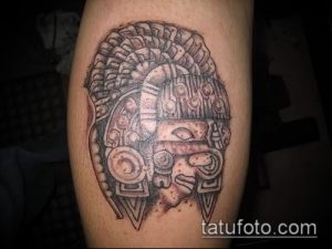 Фото тату ацтекской маски на икре, выполненное в технике blackwork. Детализированный рисунок с изображением лица в центре.