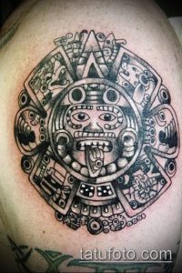 Фото тату календаря ацтеков на плече, выполненное в технике blackwork. Детализированный рисунок с изображением лица в центре.