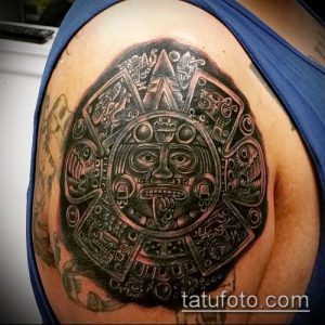 Фото тату календаря ацтеков на плече, выполненное в технике blackwork. Детализированный рисунок с изображением лица в центре.