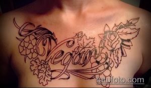 Фото тату: надпись "Vegan" в окружении винограда, листьев и цветов, выполненная в стиле лайнарт на груди. Детализированный рисунок татуировки.